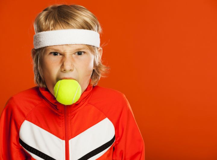boy in tennis gear with tennis ball in his mouth and angry expression